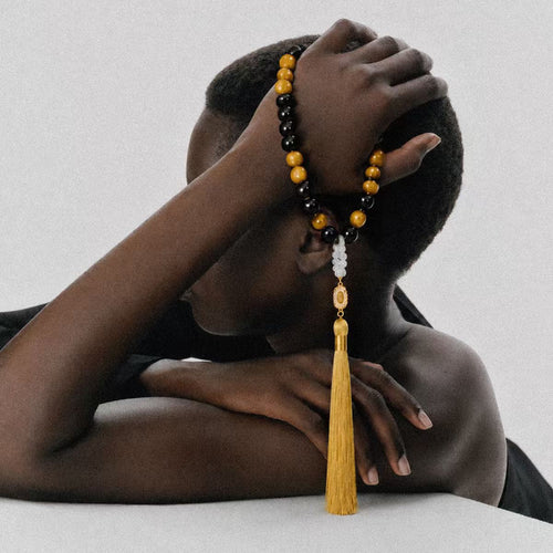 Person holding Clarity + Action Meditation Bracelet which is a black and yellow beaded bracelet with a gold tassel against a neutral background