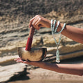 Hand holding a singing bowl with a wooden stick, wearing the Ease + Release Meditation Bracelet a beaded bracelet, against a natural background.