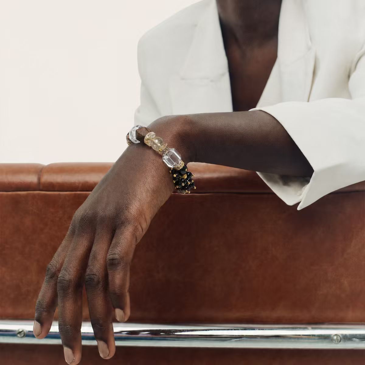 The Gold | Edge Bracelet shown on a model's wrist resting on a brown leather chair, emphasizing the contrast between the black onyx beads and the white blazer.