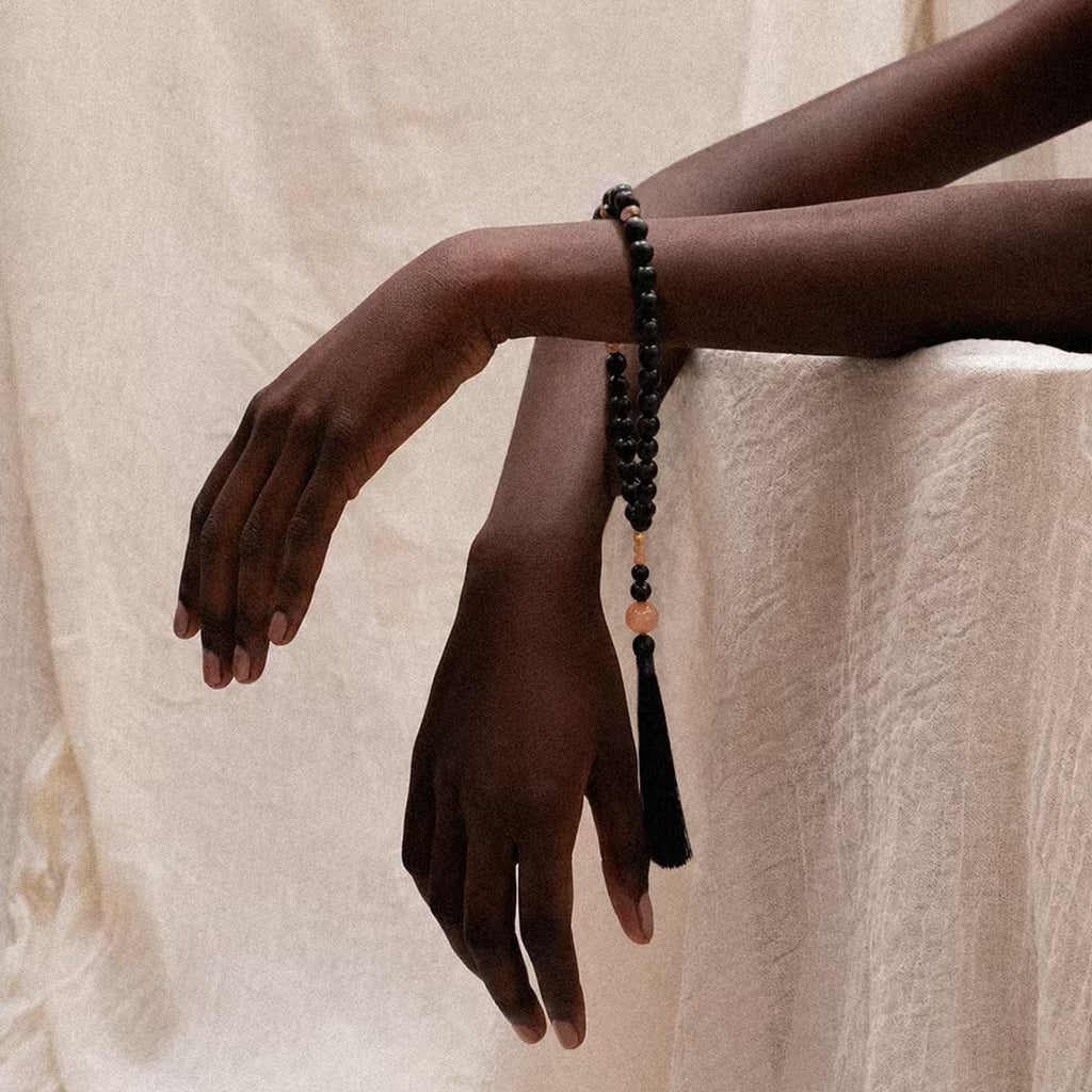 Person wearing the Release + Recovery Meditation Bracelet, a black beaded bracelet with tassel on a beige background