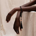 Person wearing the Release + Recovery Meditation Bracelet, a black beaded bracelet with tassel on a beige background
