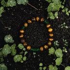 The Sumatran Tiger Crystal Bracelet by Soulucky, featuring golden-brown tiger's eye beads and textured green gemstone accents, resting on dark soil surrounded by small green leaves.