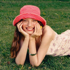A model wearing the Wood | Align handcrafted bead bracelet, styled with a pink bucket hat and floral dress while lying in a grassy field.