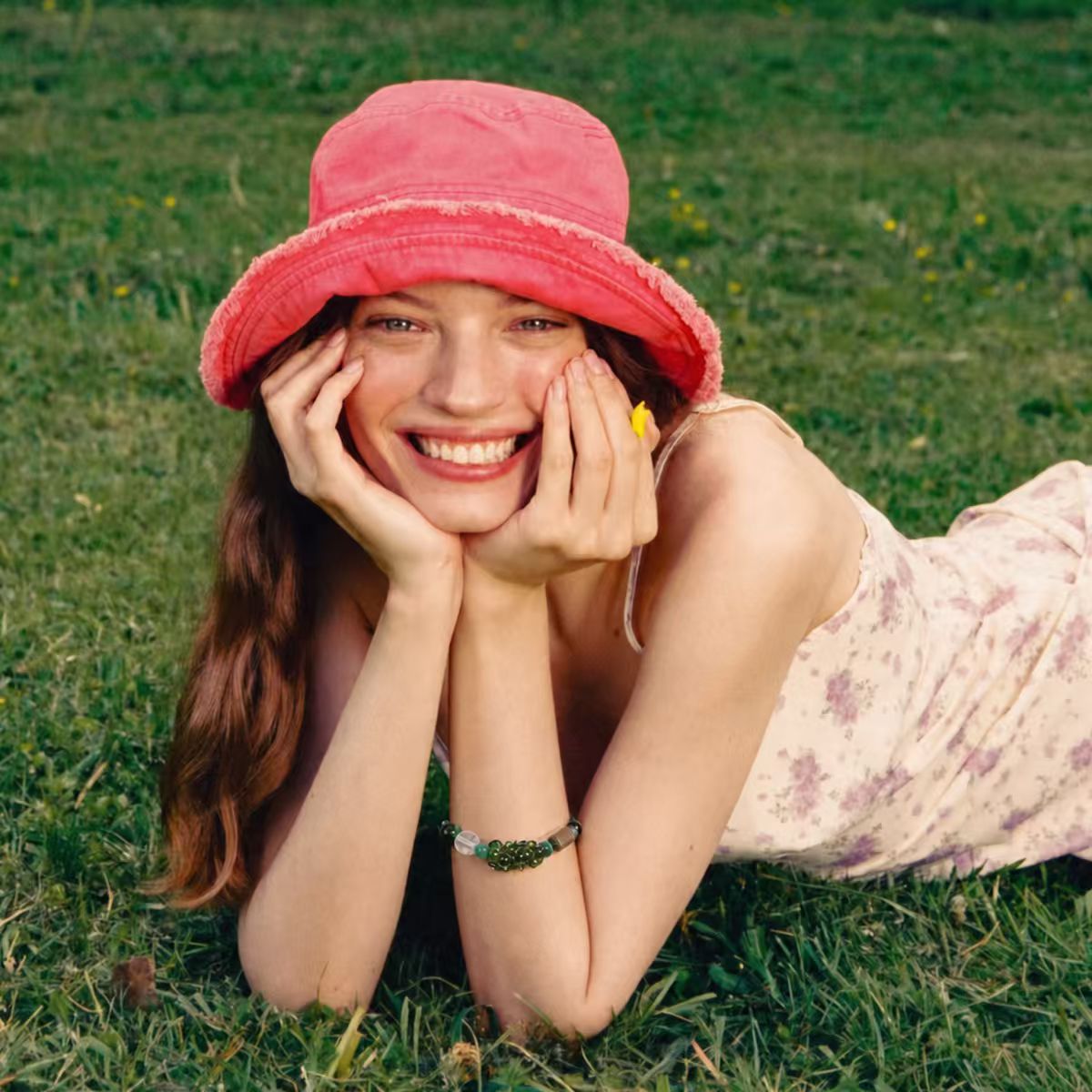 A model wearing the Wood | Align handcrafted bead bracelet, styled with a pink bucket hat and floral dress while lying in a grassy field.
